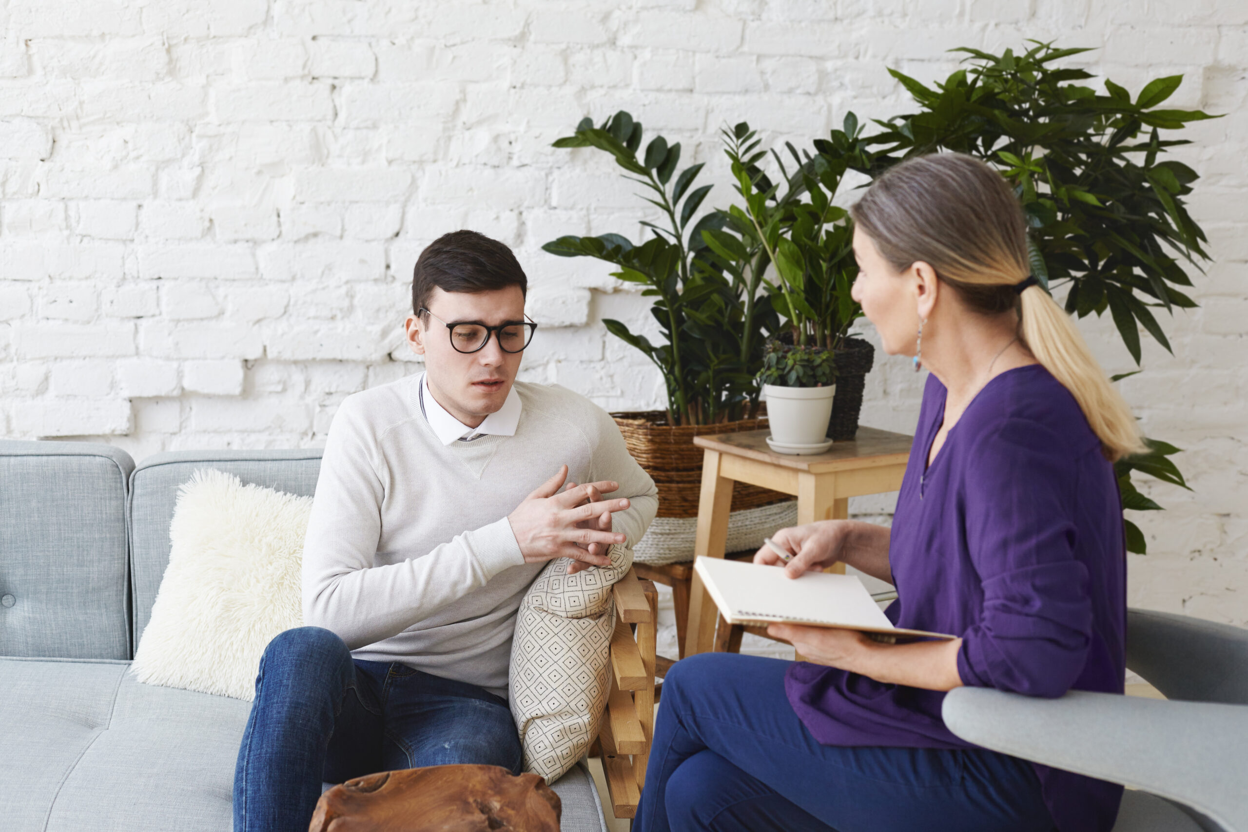 candid shot of young man in glasses talking about his problems during psychological therapy session, sitting on coach while mature female psychologist with copybook listening to him and making notes